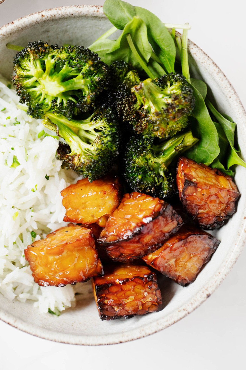 A zoomed in image of a white serving bowl, which has been filled with cooked rice, crispy roasted broccoli, spinach, and a plant based protein.