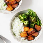 Two ceramic bowls hold baked lemon pepper tempeh cubes, roasted broccoli, and white rice. Two serving forks are resting nearby.