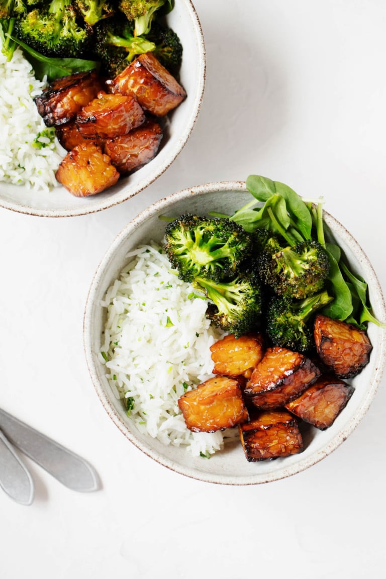 Two ceramic bowls hold baked lemon pepper tempeh cubes, roasted broccoli, and white rice. Two serving forks are resting nearby.