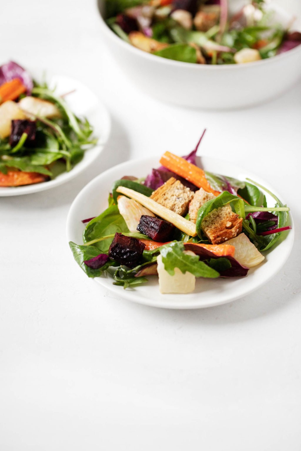 An angled photo of small salad plates, topped with roasted root vegetables, greens, and cubed bread.