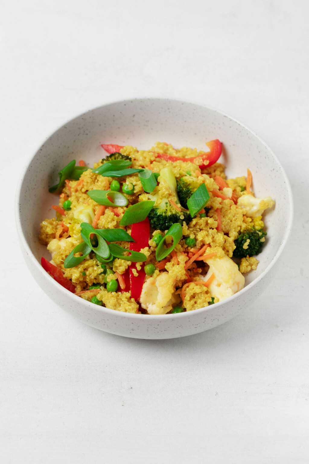 A round, white bowl holds a golden mixture of quinoa and vegetables, which has been topped with green onions and pieces of chopped cashew nuts.