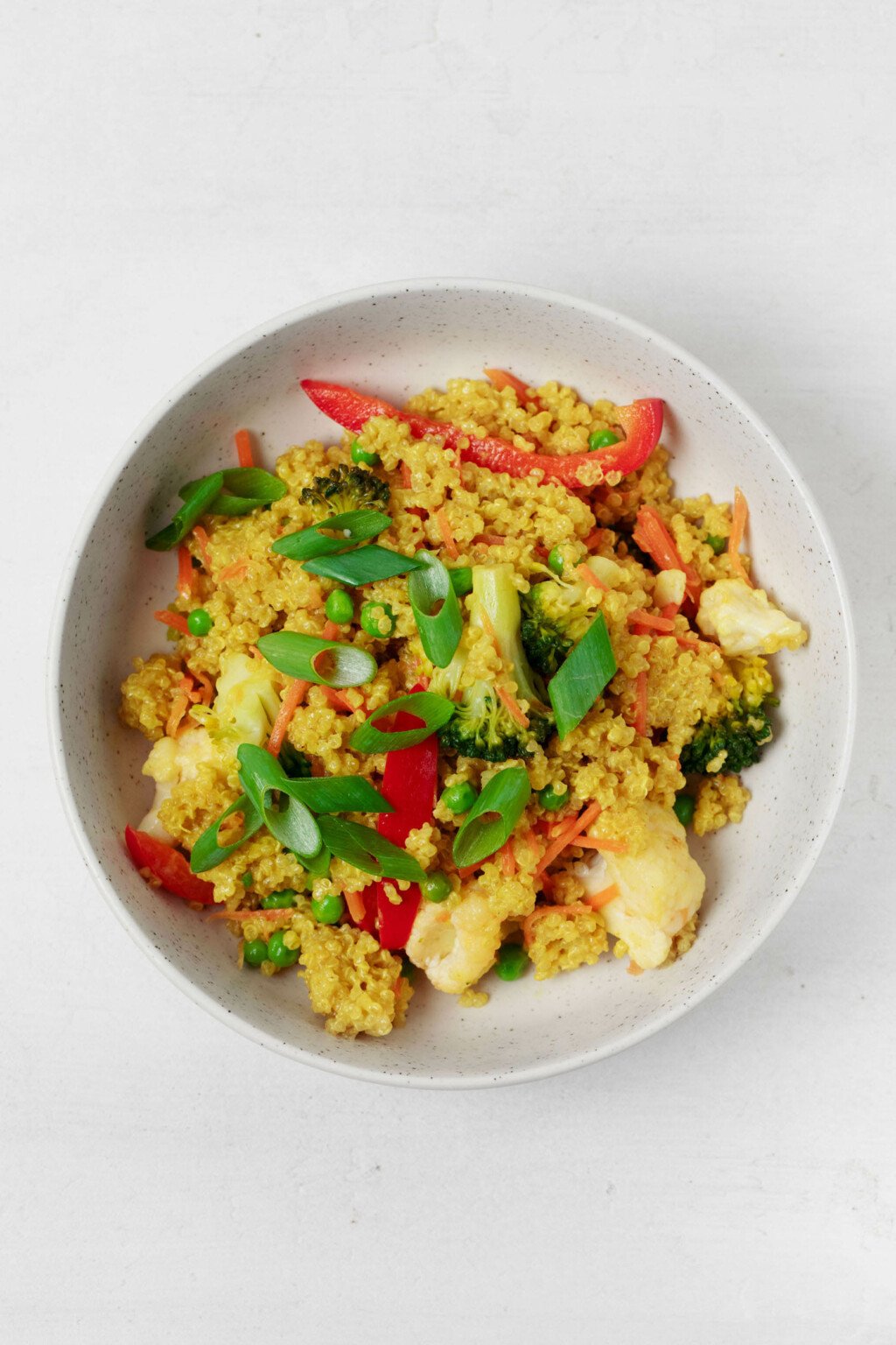 An overhead, birds eye image of a bowl of quinoa and vegetables, which have been topped with green onions. The bowl rests on a white surface.