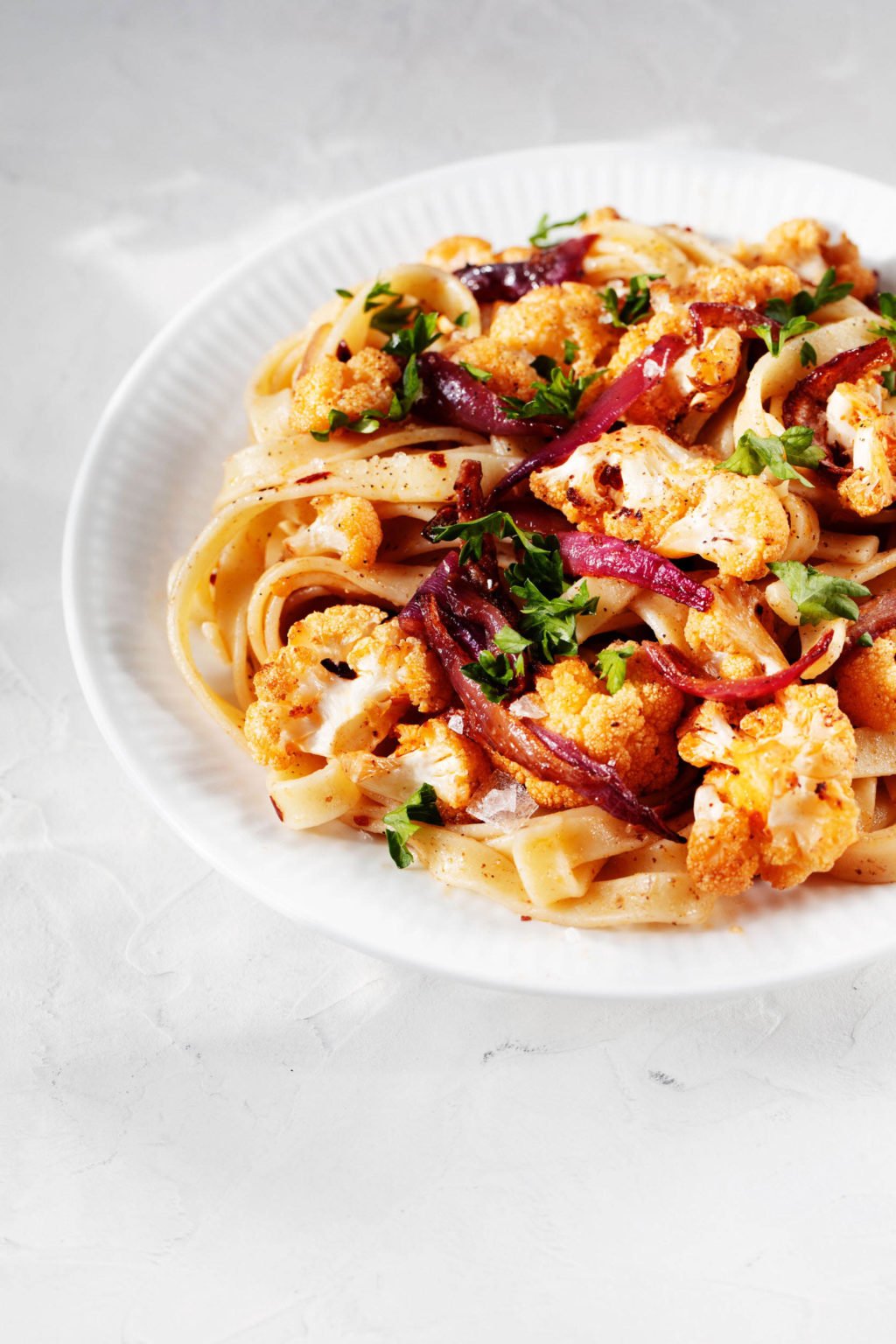 An angled image of a dish of roasted cauliflower pasta. It is resting on a white rimmed plate on a white surface.