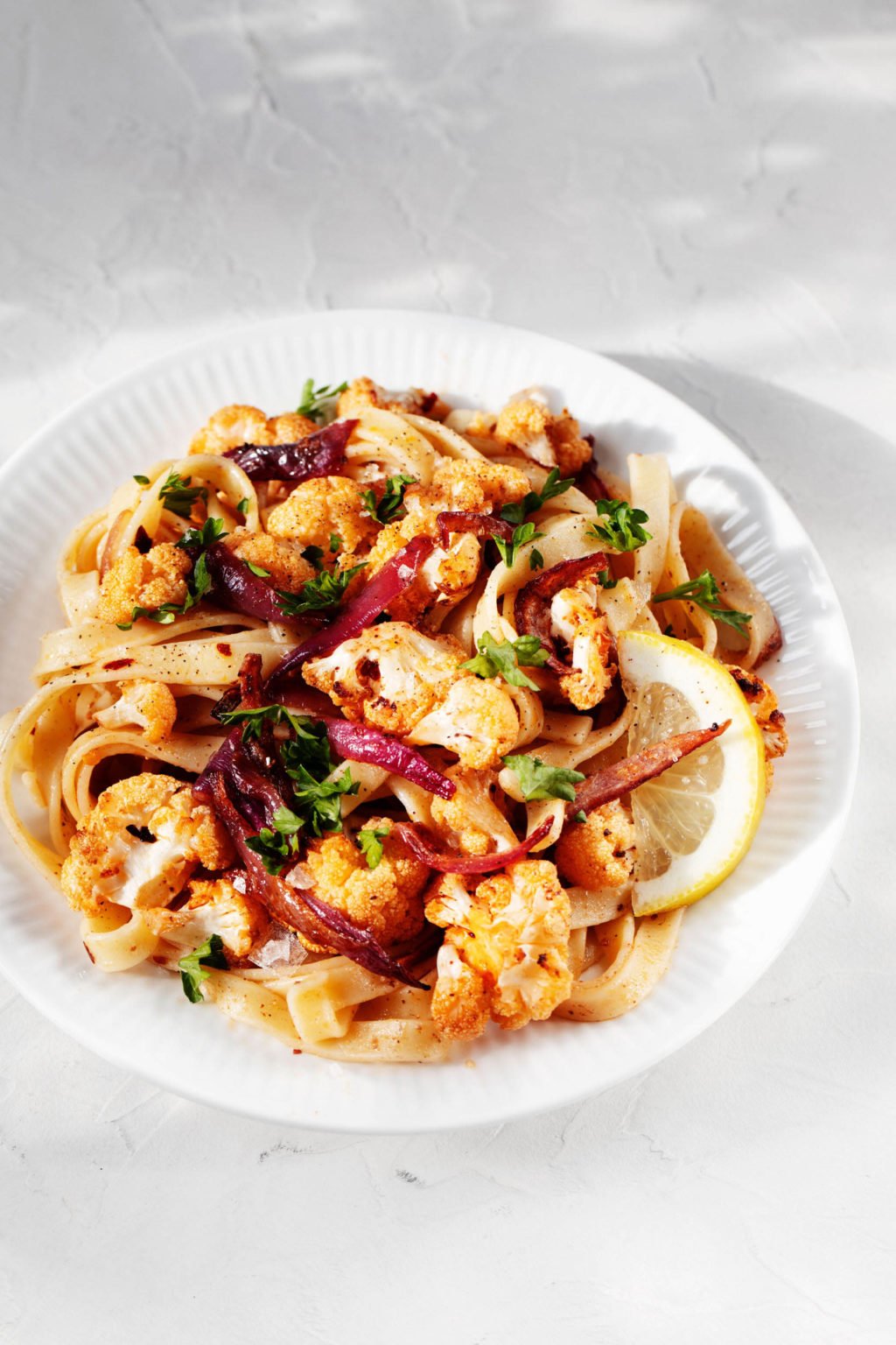 A plate of pasta with roasted cauliflower florets and red onion is served on a white rimmed plate. It rests on a white surface.