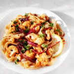 A plate of pasta with roasted cauliflower florets and red onion is served on a white rimmed plate. It rests on a white surface.