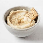 A small ceramic bowl holds a creamy vegan cashew cheese and a cracker for dipping.