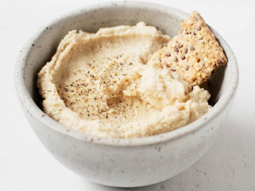 A small ceramic bowl holds a creamy vegan cashew cheese and a cracker for dipping.