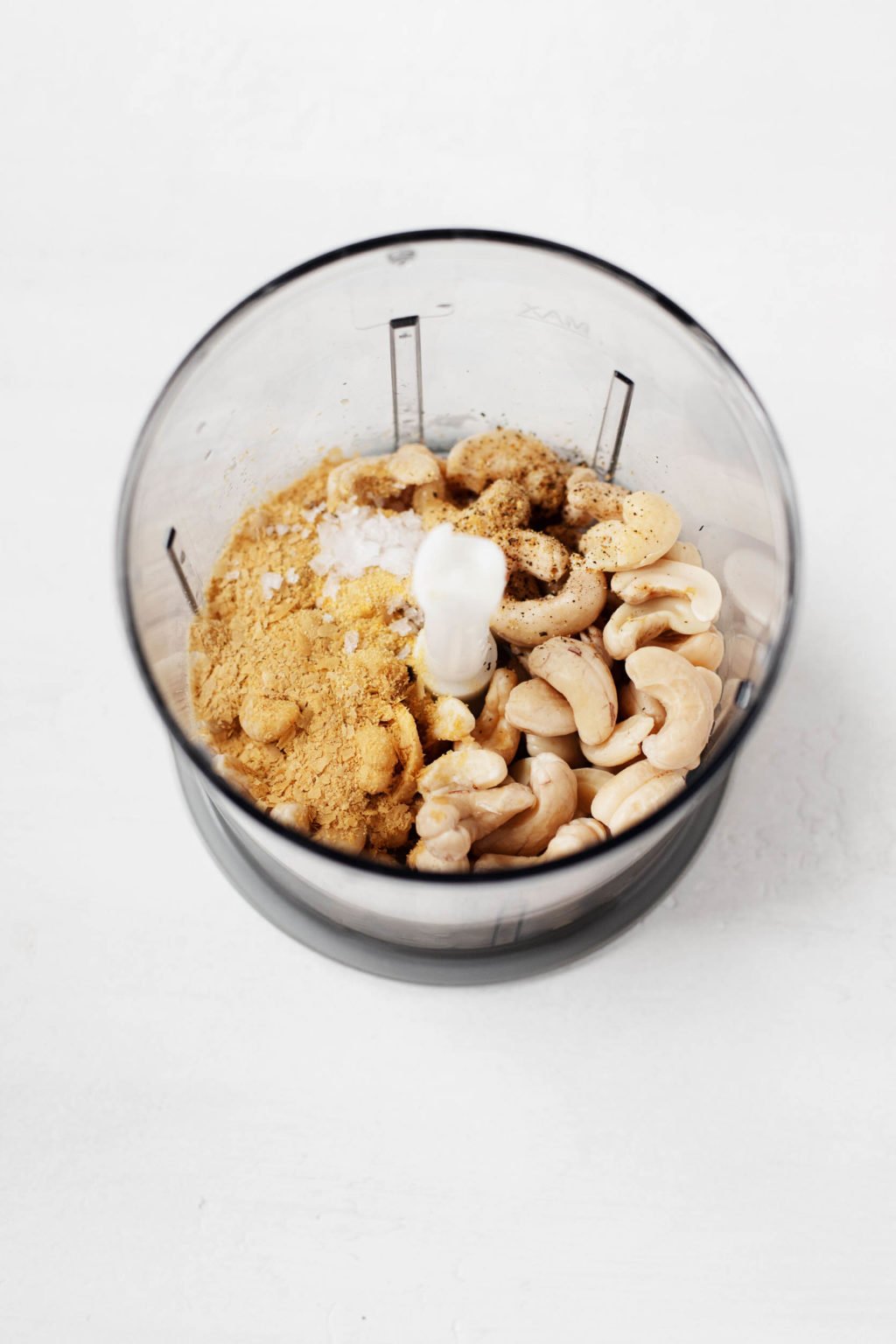 A food processor is full of raw cashews, nutritional yeast, and salt, ready for blending.