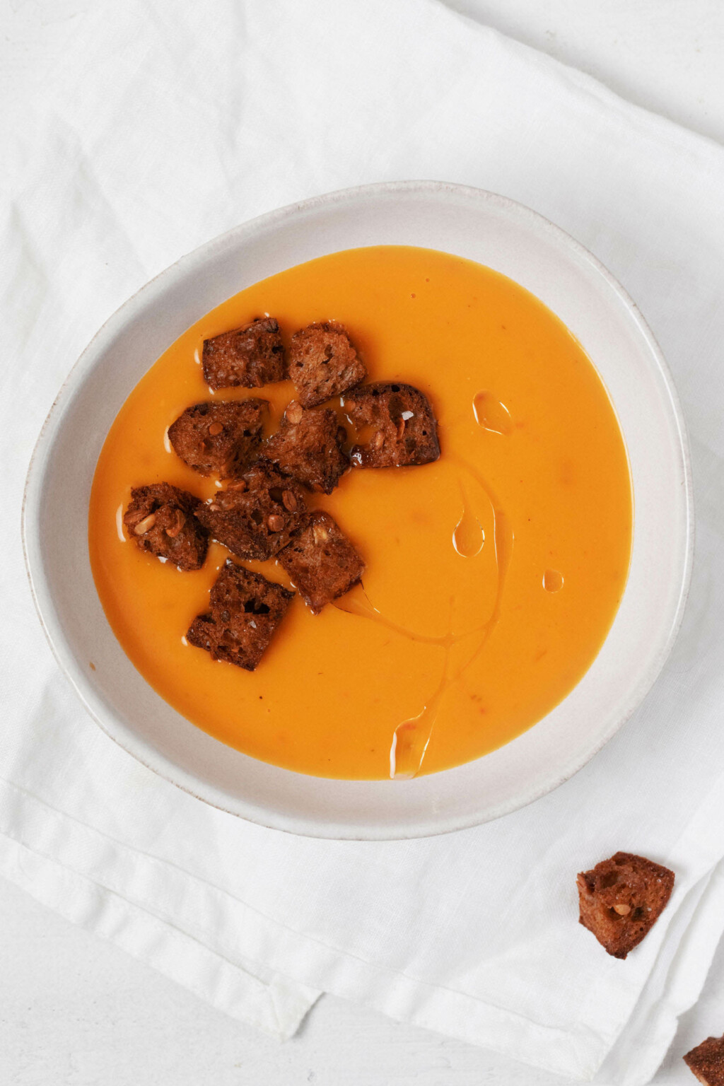 An asymmetrical white bowl holds a creamy vegan butternut squash soup that is seasoned with five spice powder. It's topped with croutons.