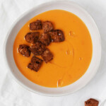 An asymmetrical white bowl holds a creamy vegan butternut squash soup that is seasoned with five spice powder. It's topped with croutons.