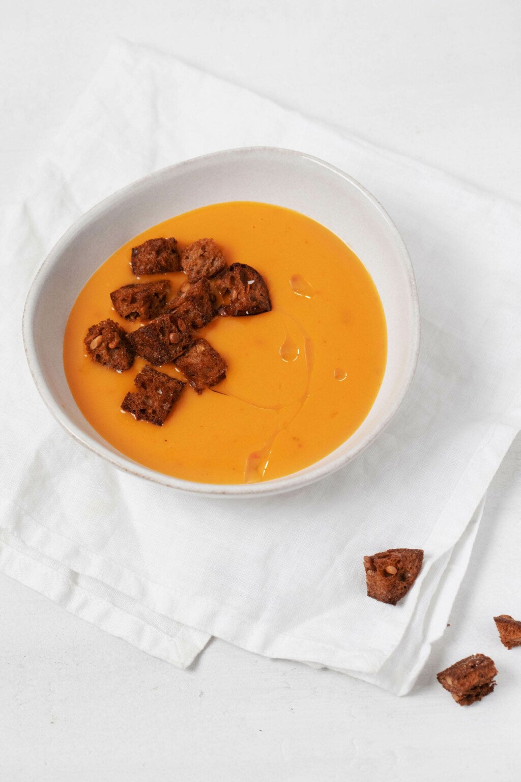 A bowl of orange hued soup has been topped with dark brown, crispy croutons.