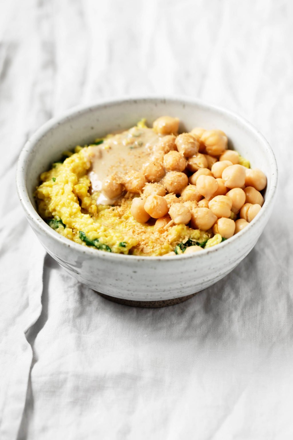 A gray and white ceramic bowl holds a dish of savory oatmeal made with chickpeas and spinach. It has been topped with a spoonful of tahini.