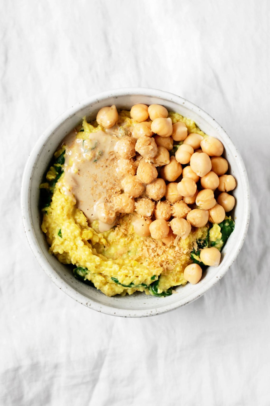 A bowl containing savory, turmeric spiced oats with chickpeas and greens is on a gray linen cloth.