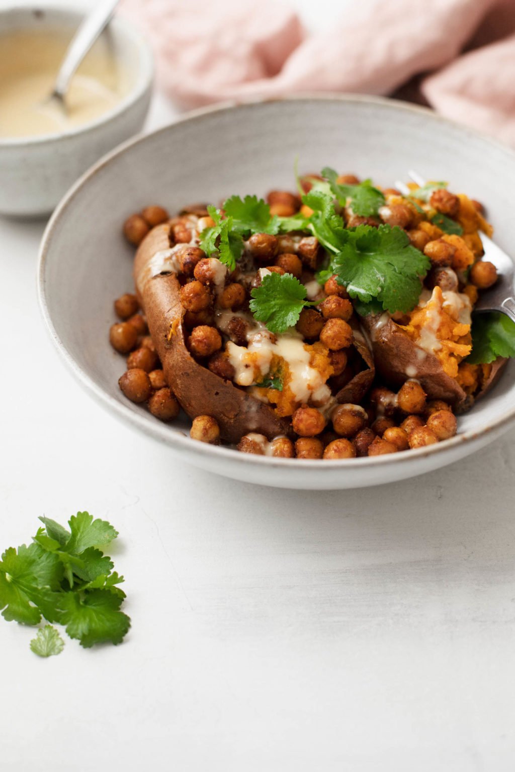 A small bowl holds a baked sweet potato that's been stuffed with spiced chickpeas, herbs, and sauce.