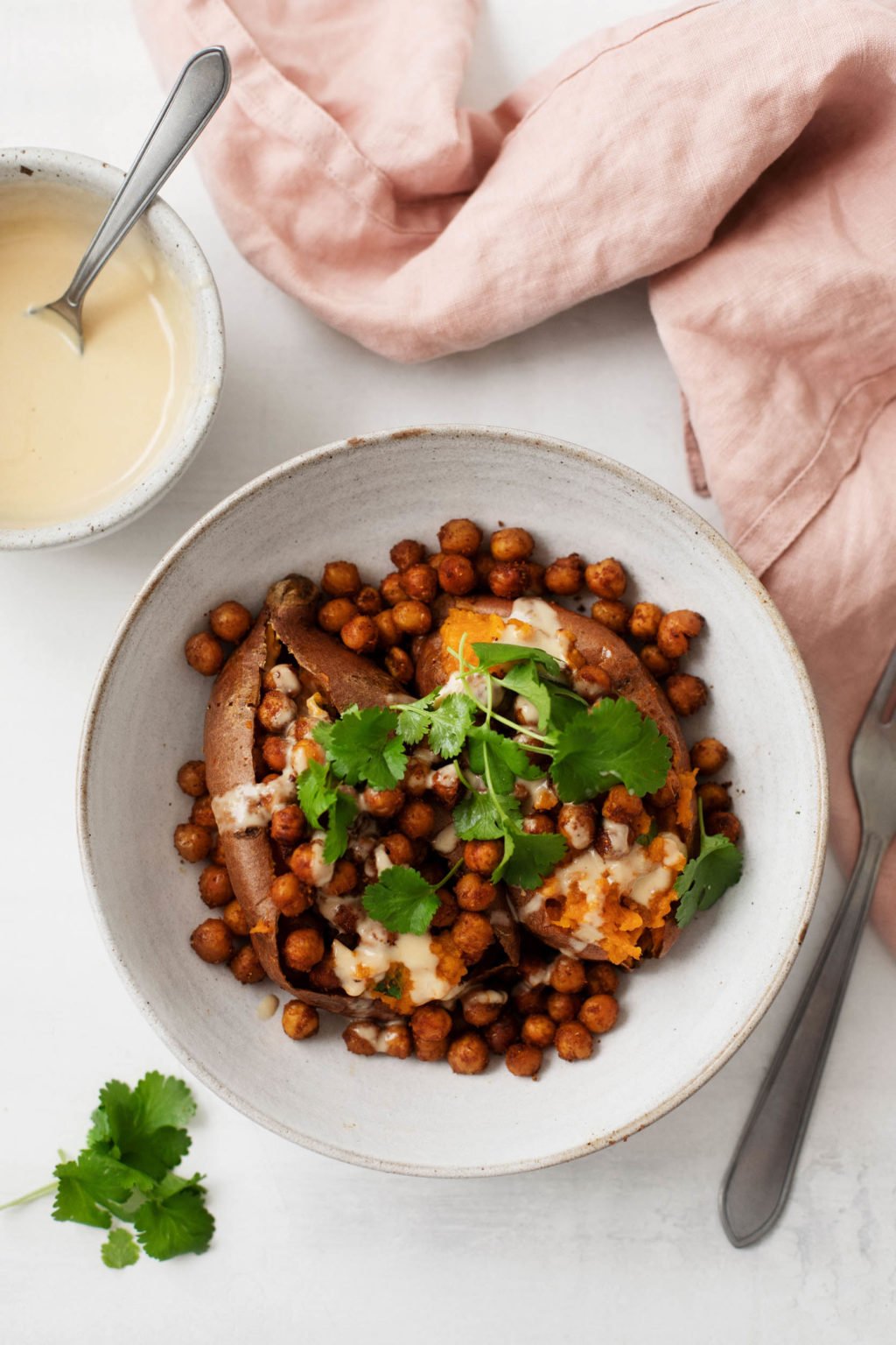 A bowl of baked potatoes and roasted chickpeas, topped with cilantro and served with a small bowl of sauce.