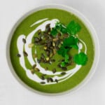 A bowl of vegan green soup has been garnished and plated in a white bowl. The bowl rests on a white surface.