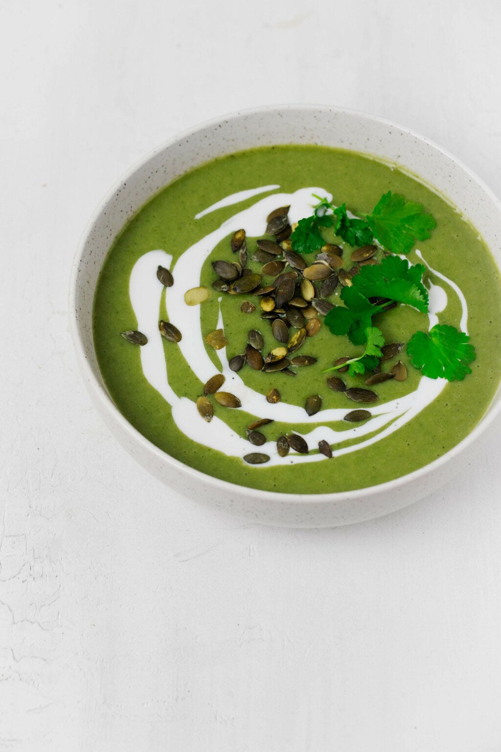 A bowl of soup, garnished with herbs and pepitas, is resting on a white surface.
