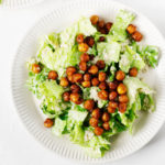 Two white, fluted ceramic plates have been piled with a vegan quinoa chickpea caesar salad. The plates are resting on a white background.