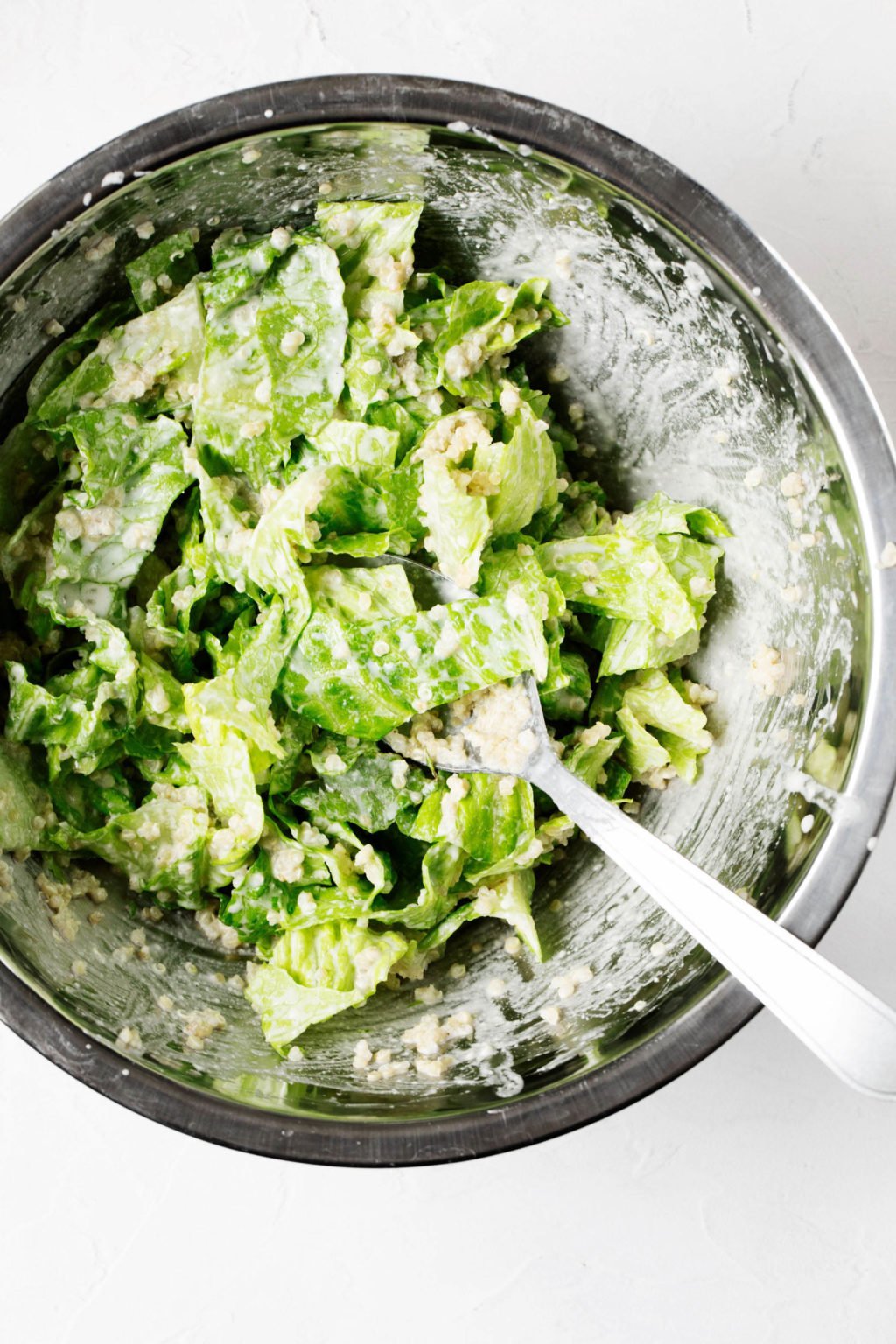A large, metal mixing bowl is filled with romaine lettuce, which is being tossed with a creamy salad dressing.