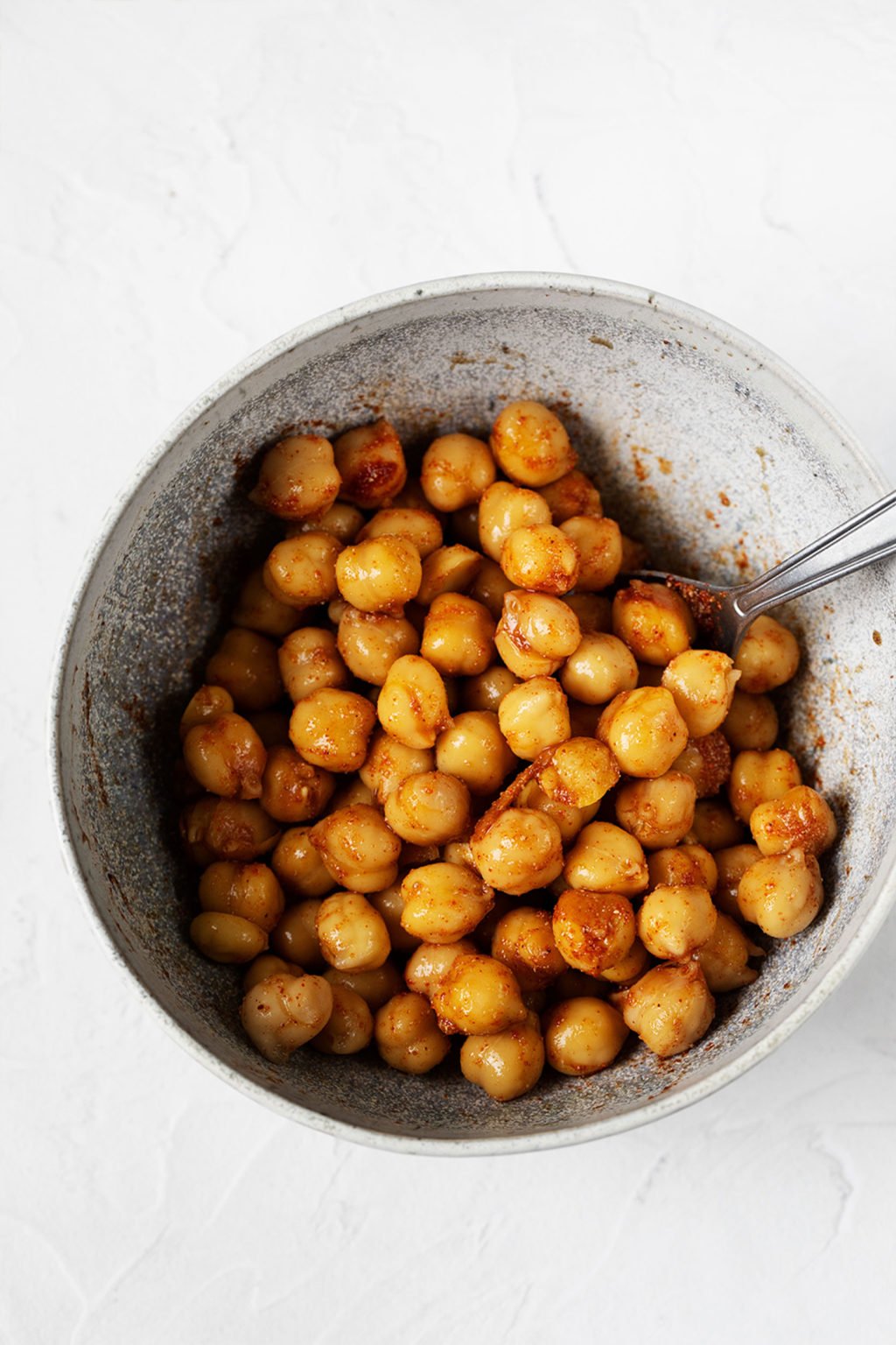 A small, white and gray ceramic bowl holds chickpeas that are being tossed with olive oil and spices.