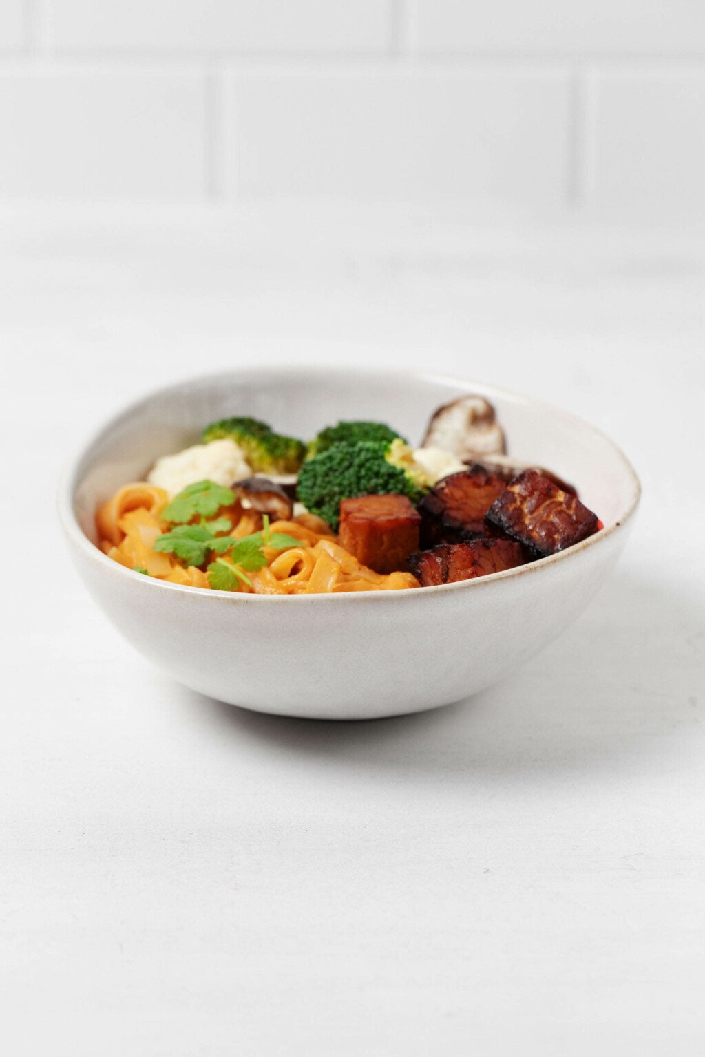 Noodles with an orange sauce, cubes of tempeh, and vegetables are piled into an asymmetrically shaped, oval bowl. It is pictured against a white surface.