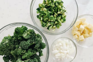 Chopped vegetables and greens are in clear, glass bowls, resting on a white surface.