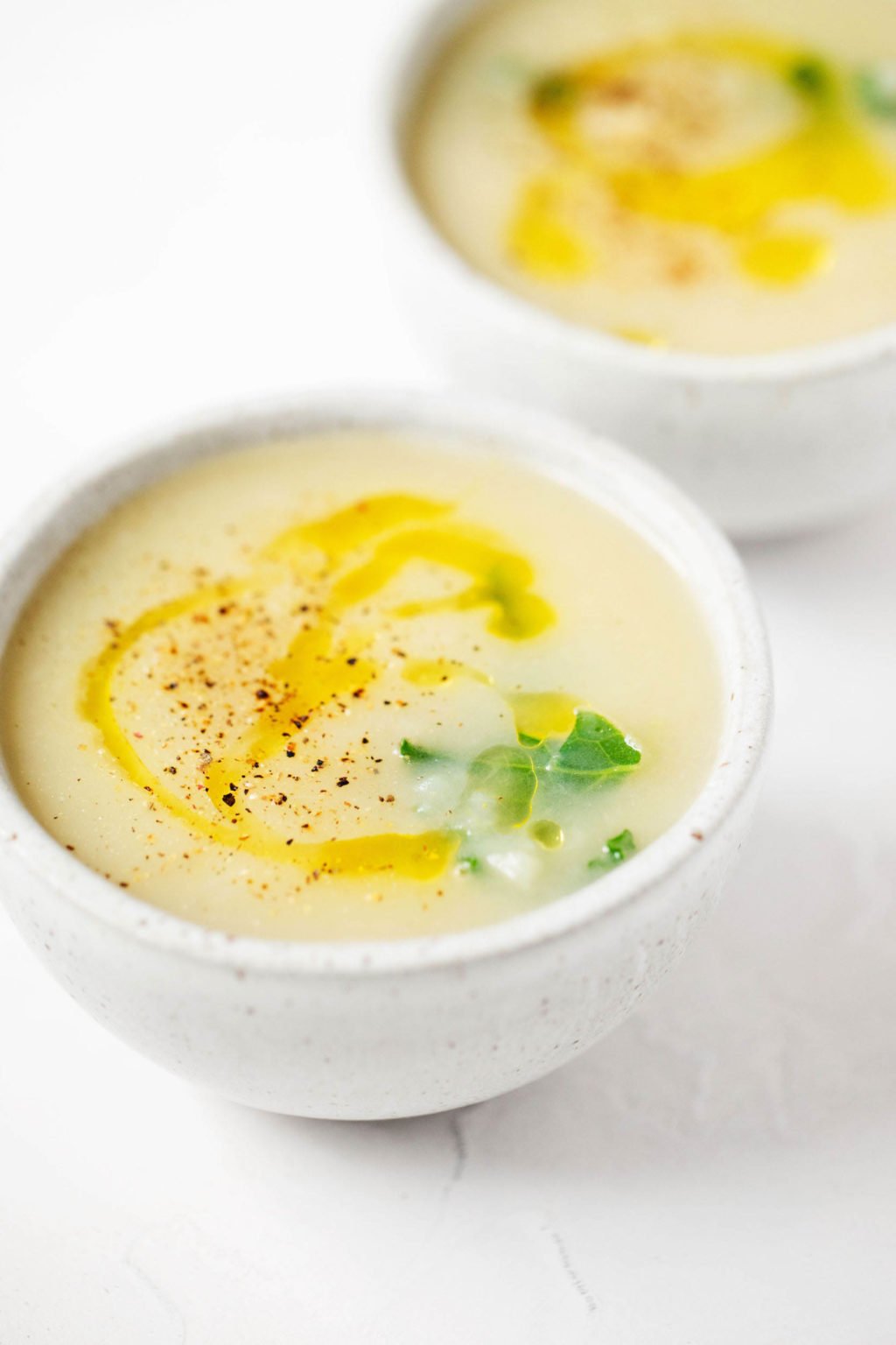 Two bowls of a plant based soup are topped with a swirl of olive oil and a turn of cracked black pepper.