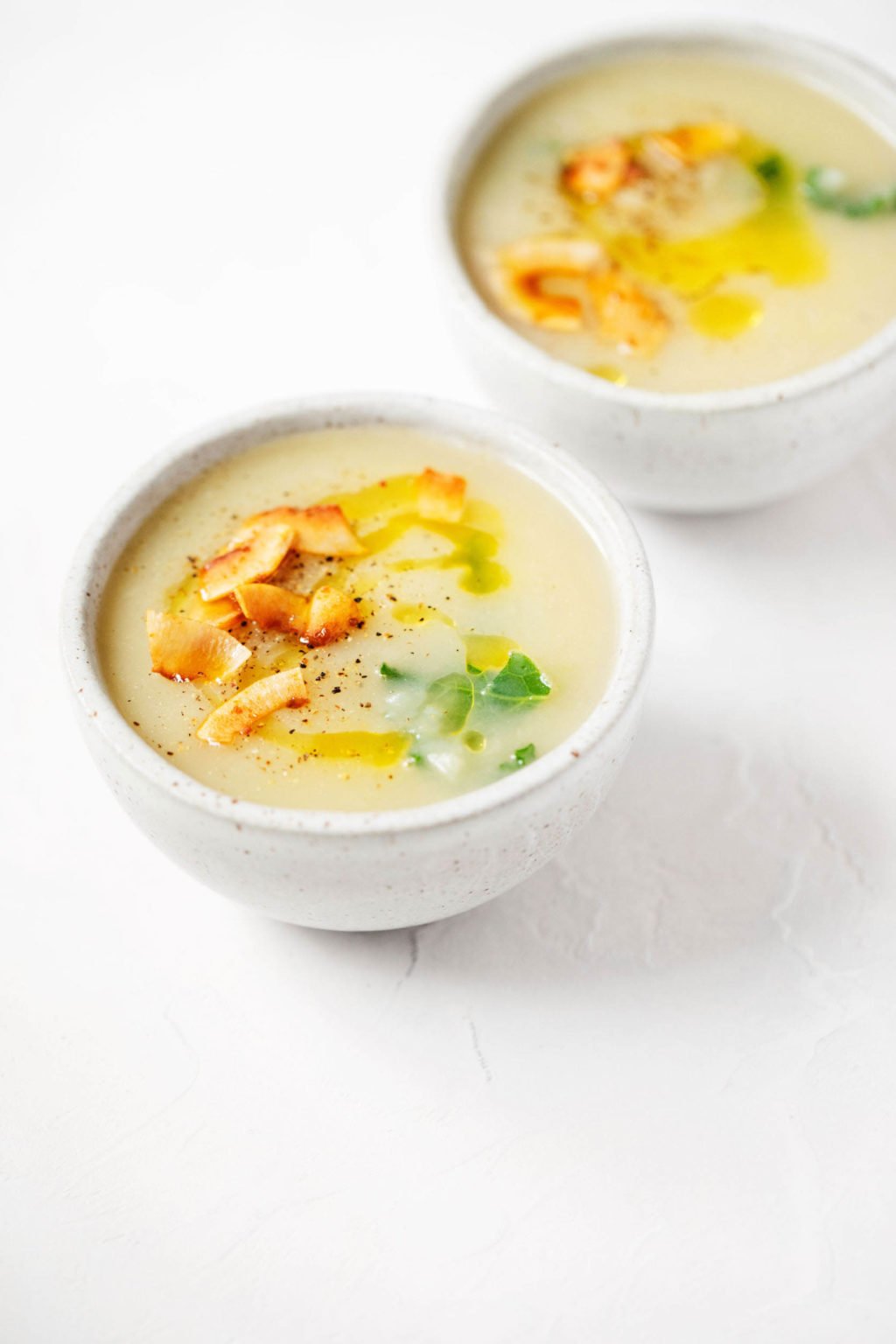 Two portions of a vegan potato leek soup are resting in small, round white soup bowls. They're laid out on a bright, white surface.