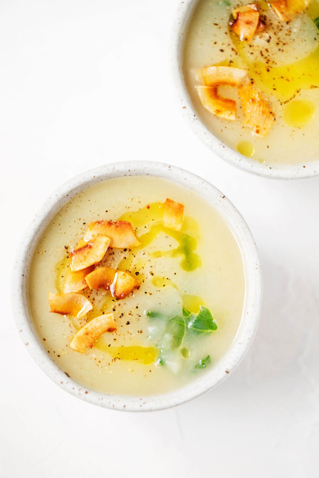 Two white bowls of a creamy vegan potato leek soup rest on a white surface. They're topped with coconut bacon and greens.