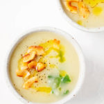 Two white bowls of a creamy vegan potato leek soup rest on a white surface. They're topped with coconut bacon and greens.
