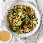 A dish of cabbage, broccoli, whole grains, and legumes has been plated in a ceramic bowl that's resting on a light gray colored cloth. There's a small bowl of peanut sauce near the bowl.