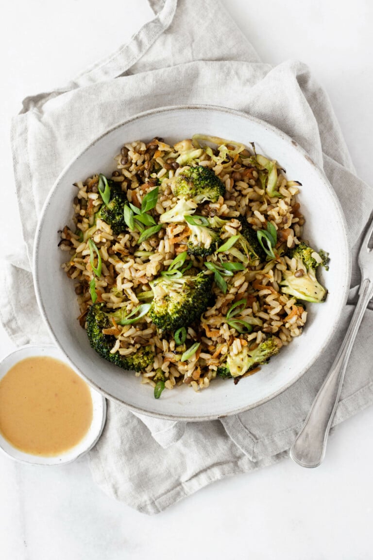 A dish of cabbage, broccoli, whole grains, and legumes has been plated in a ceramic bowl that's resting on a light gray colored cloth. There's a small bowl of peanut sauce near the bowl.