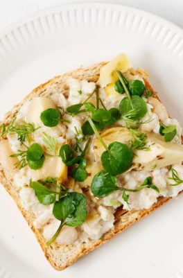 A slice of toast is topped with smashed white bean salad, artichokes, and microgreens.