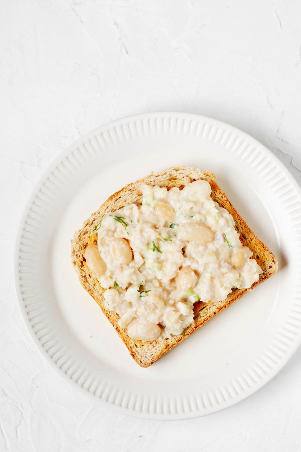 A piece of toast, topped with legumes, is resting on a round, rimmed white plate. It sits on a white surface.