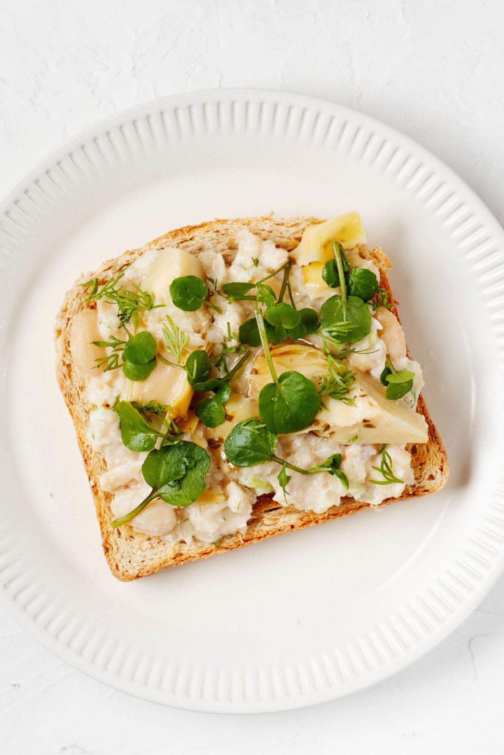An overhead image of a piece of toast with white bean salad and microgreens. It sits on a white, rimmed plate upon a white surface.