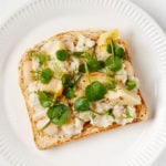 An overhead image of a piece of toast with white bean salad and microgreens. It sits on a white, rimmed plate upon a white surface.