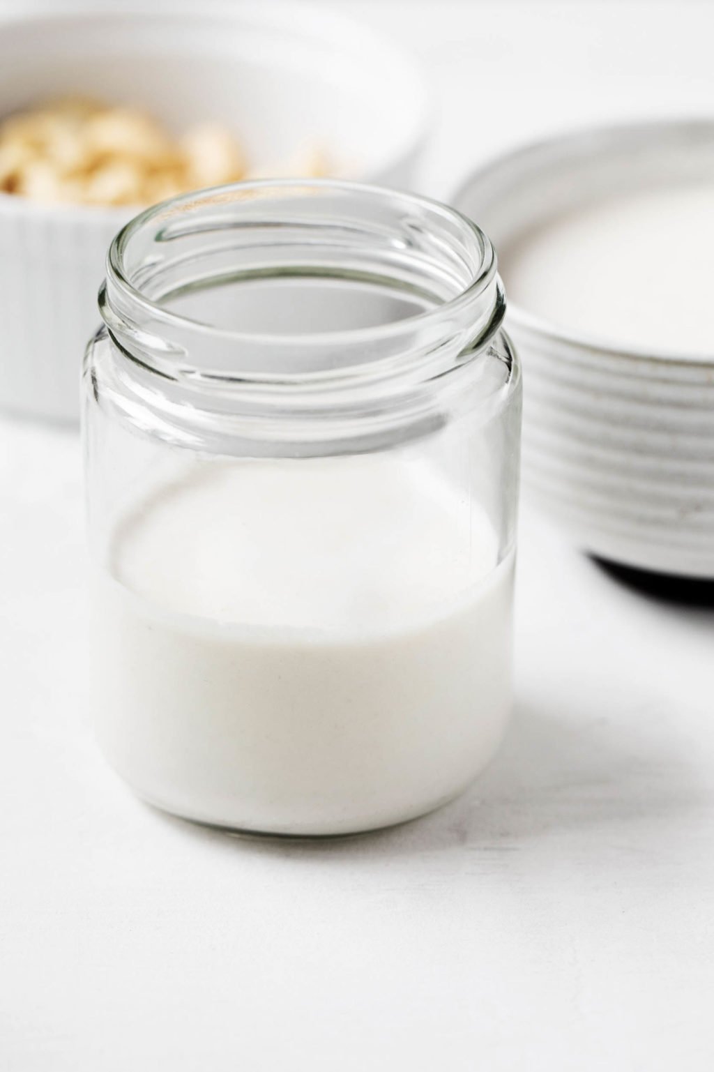 A glass jar has been filled with dairy-free cashew cream. There's another small bowl of the cream and some soaked cashews in the background.