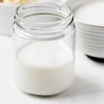 A glass jar has been filled with dairy-free cashew cream. There's another small bowl of the cream and some soaked cashews in the background.