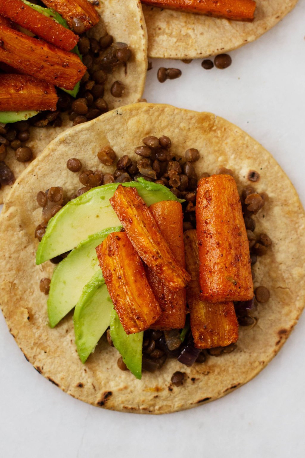 A close up, overhead shot of plant-based tacos with roasted carrots, avocado slices, and spiced lentils.