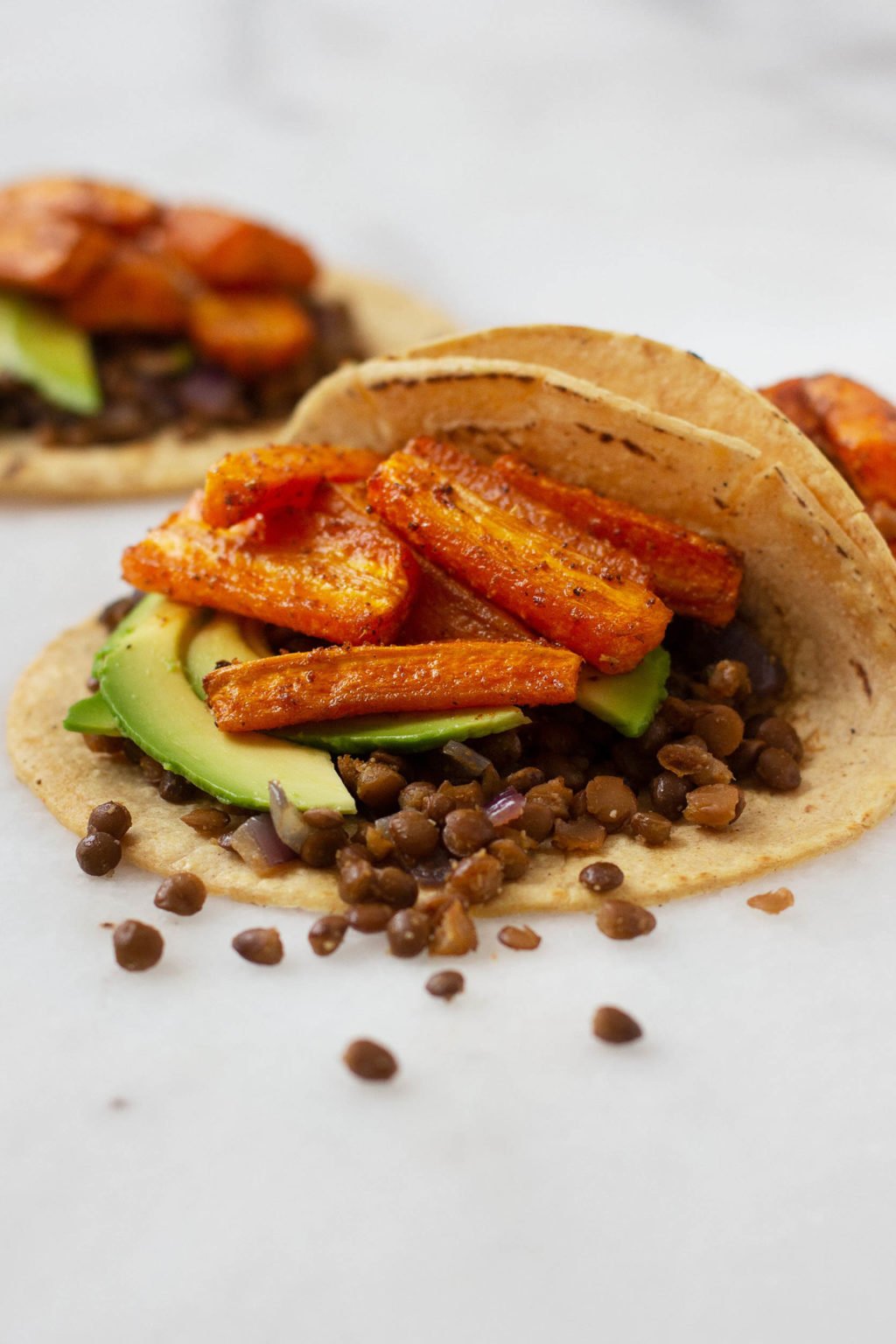 An angled shot of open, vegan tacos with a carrot and lentil filling, spilling onto a marble surface.