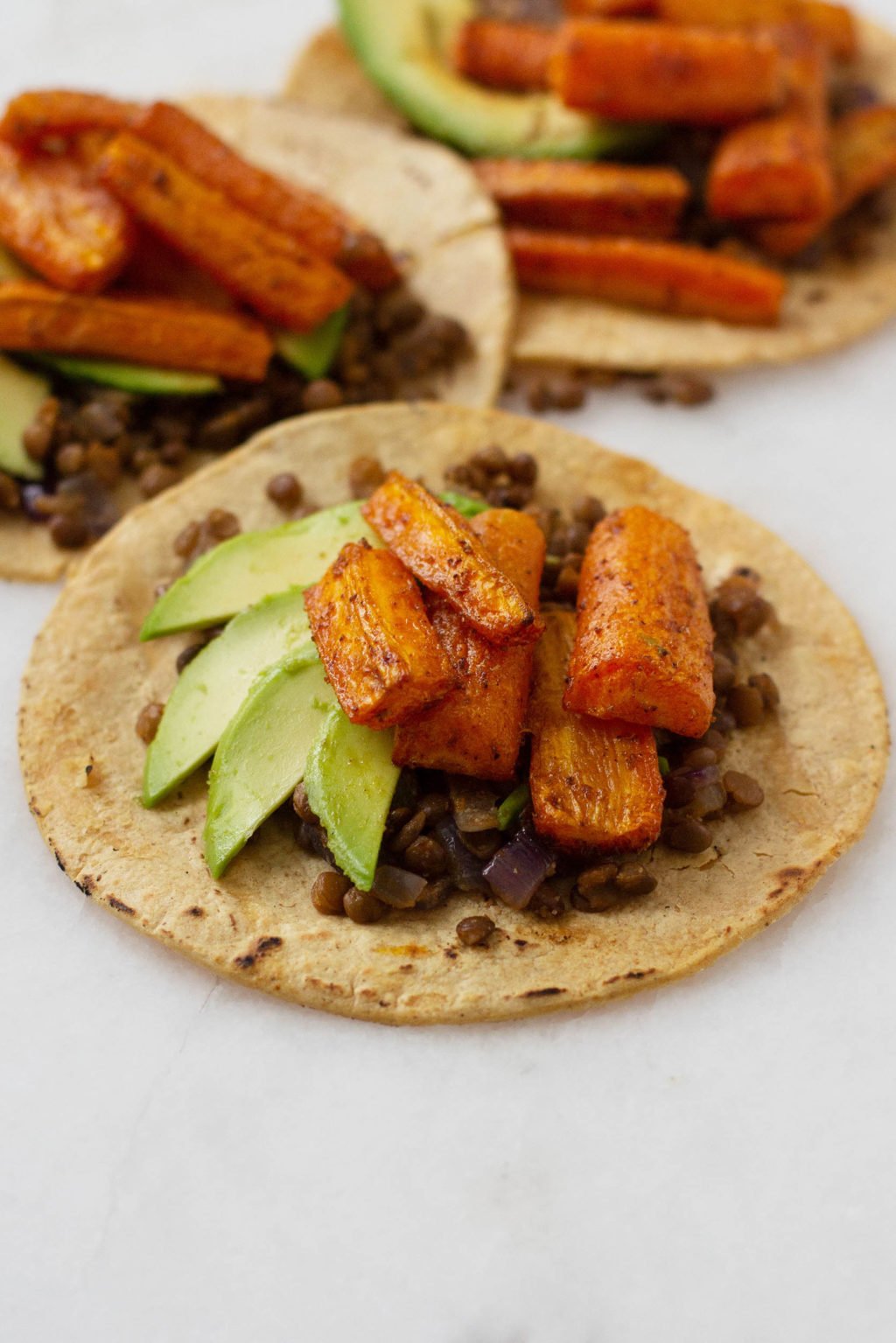 Three vegan tacos, each filled with roasted carrots, a spiced lentil and onion mixture, and avocado slices.