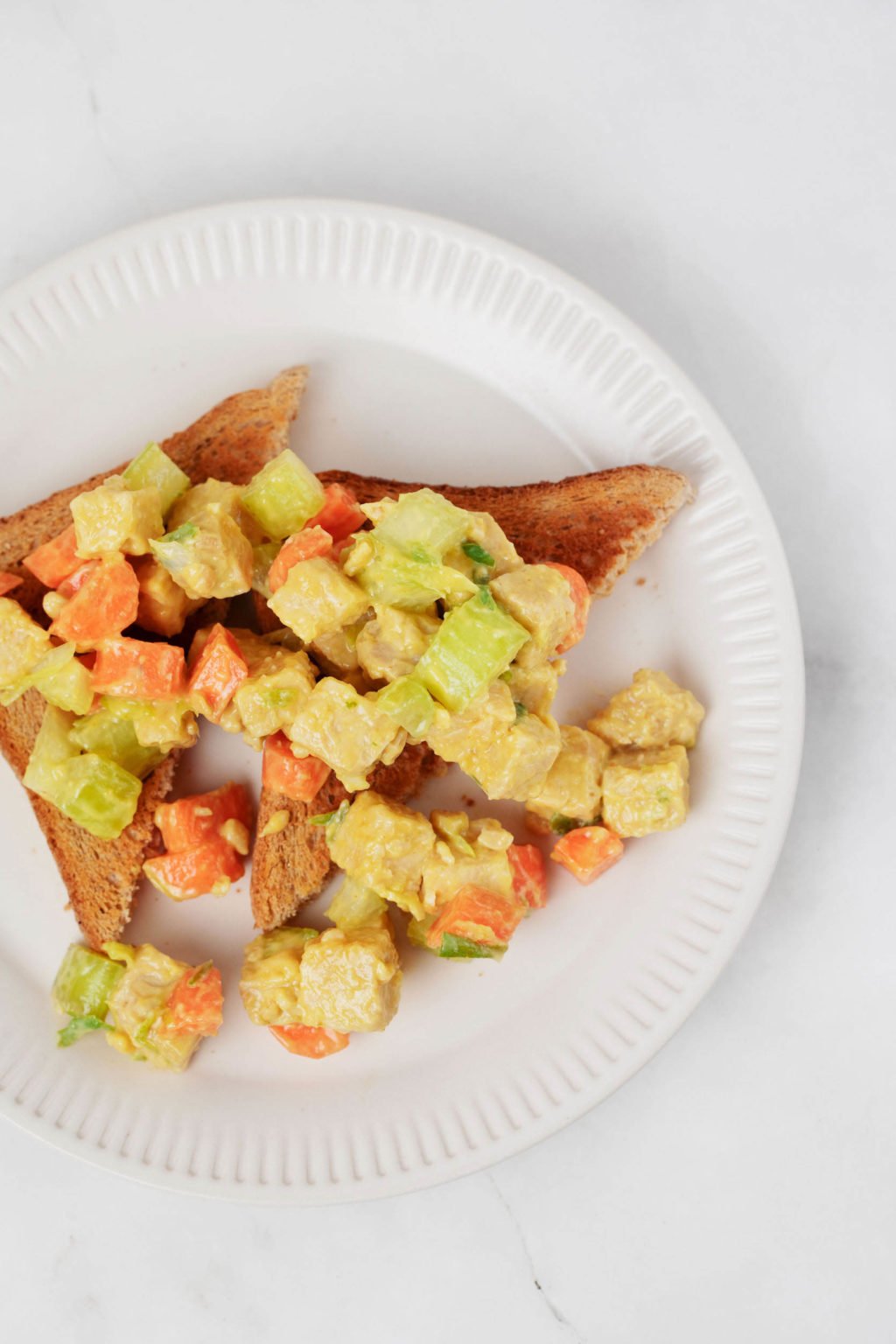 A plant-based tempeh lunch salad has been served over slices of toast, which rest on a white, round plate.