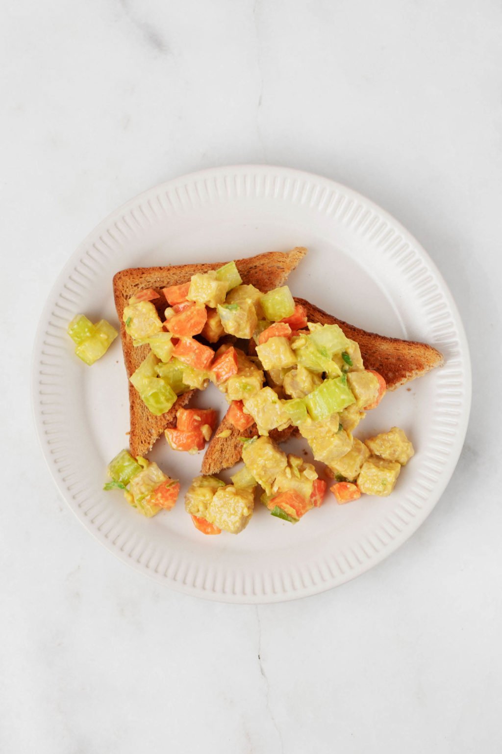 Two slices of toast have been topped with a protein and vegetables. The toast is served on a round white plate.