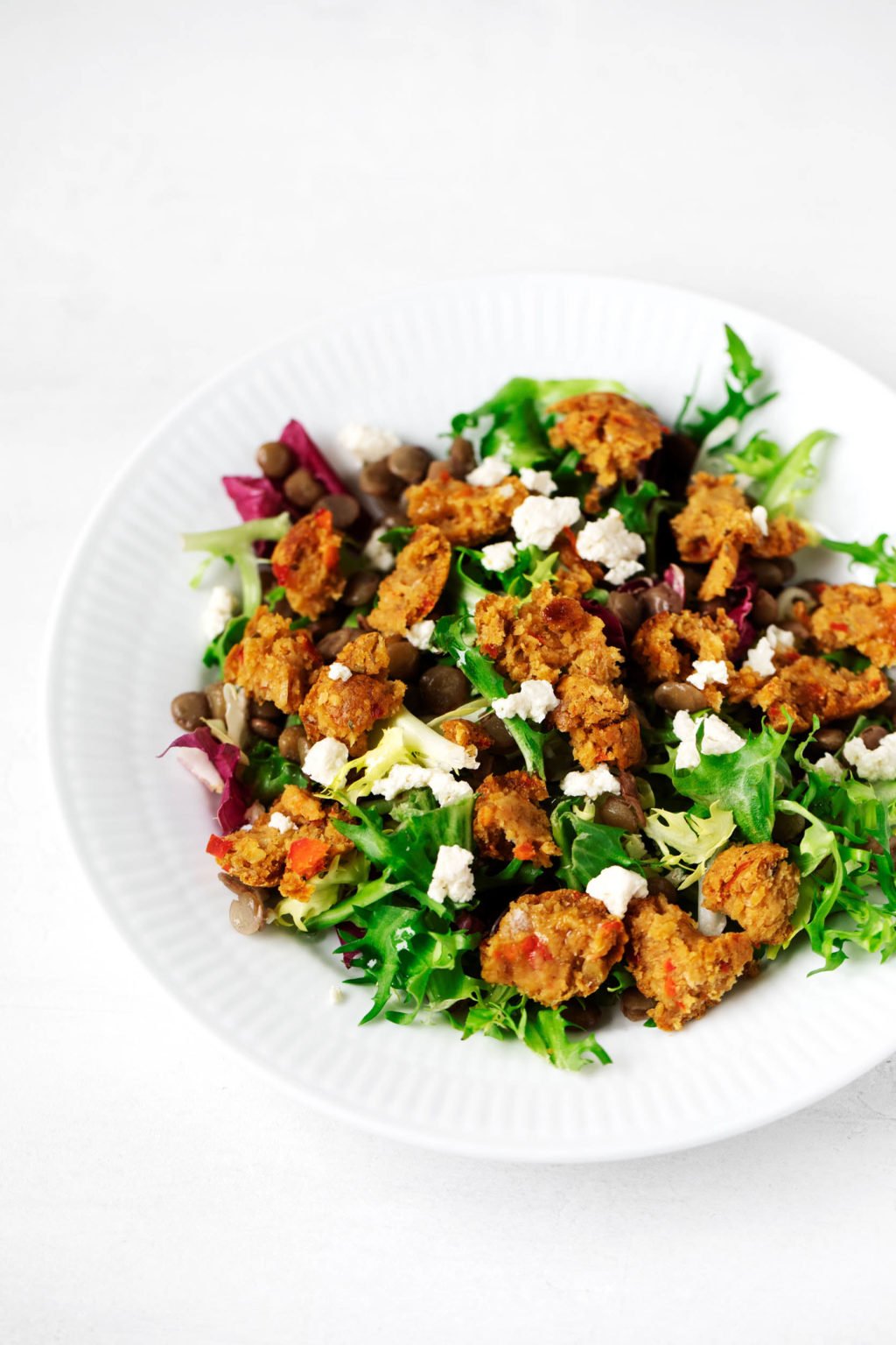 A white bowl has been filled with salad greens, vegan cheese, and savory plant-based chickpea oat balls.