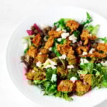 A white bowl has been filled with salad greens, vegan cheese, and savory plant-based chickpea oat balls.