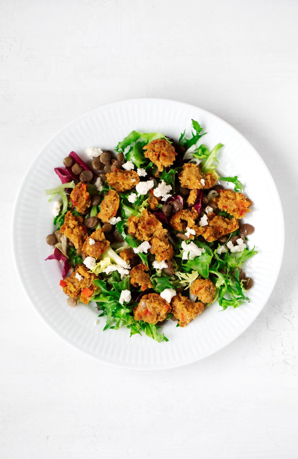 A bright, white, shallow salad bowl has been piled with greens and other plant based ingredients.