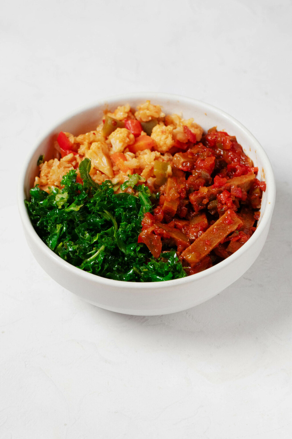An image of a round white bowl, which has been filled with a colorful arrangement of rice, vegetables, spiced seitan, and curly green kale. It rests on a white surface.