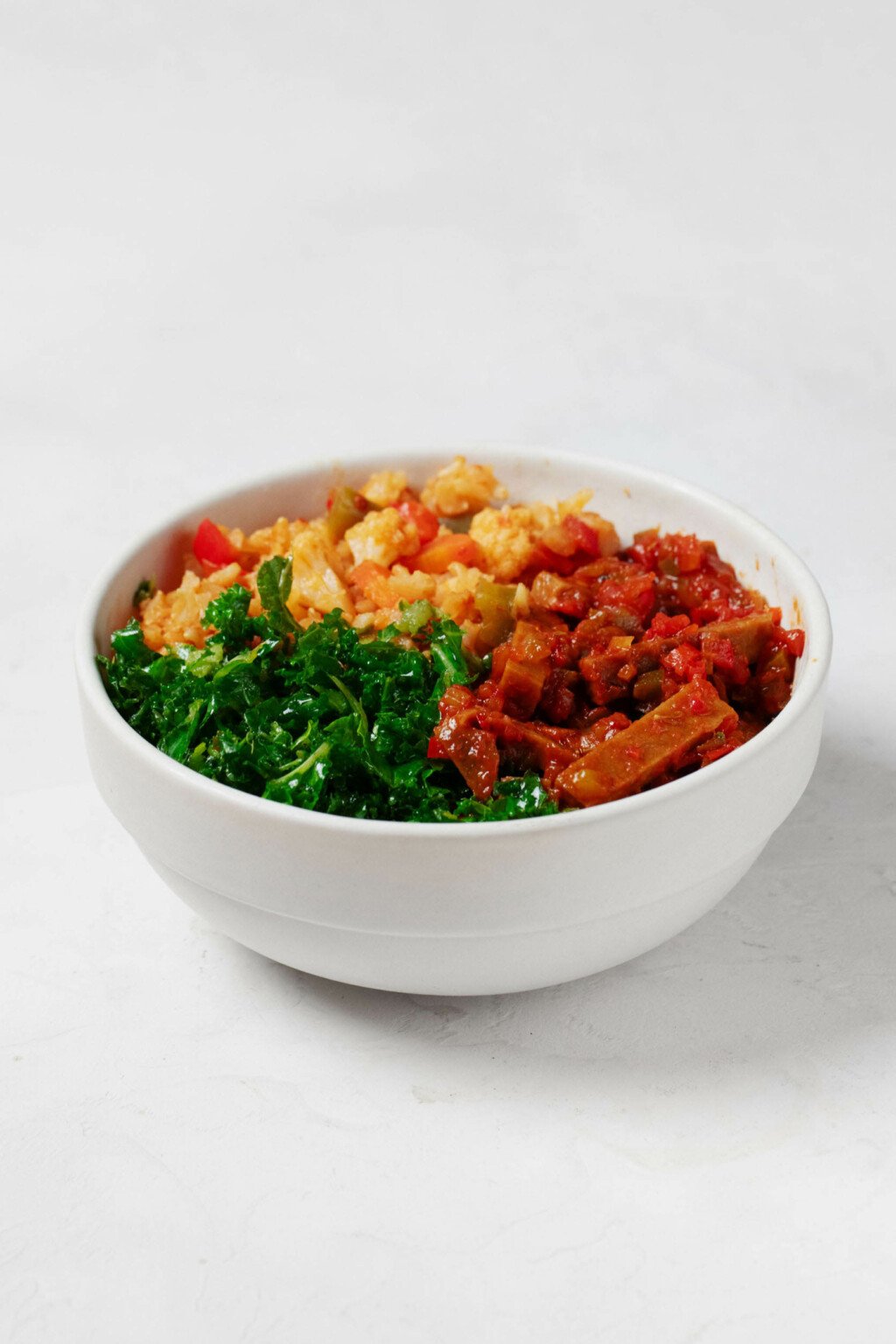 An image of a round white bowl, which has been filled with a colorful arrangement of rice, vegetables, spiced seitan, and curly green kale.
