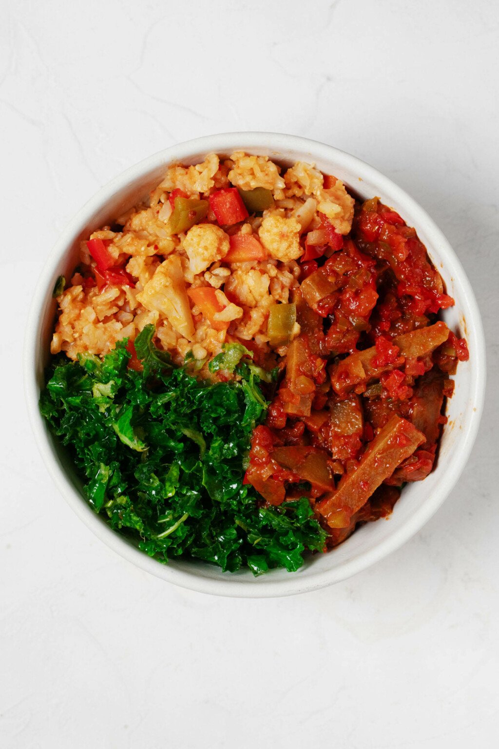 An overhead image of a round white bowl, which has been filled with a colorful arrangement of rice, vegetables, spiced seitan, and curly green kale.