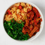 An overhead image of a round white bowl, which has been filled with a colorful arrangement of rice, vegetables, spiced seitan, and curly green kale.